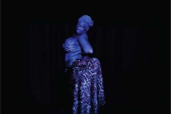 Figure 4. Silvia Sopale, lit to appear dark blue, portrays Saartjie (Sarah) Baartman. Here, her nudity conveys Baartman’s lack of agency while her gaze restores agency and humanity. Blackface y otras vergüenzas, Barcelona, 2019. (Photo by Heidi ; courtesy of Silvia Sopale)